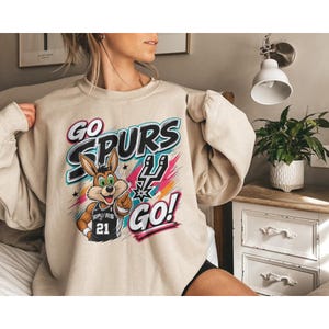May include: A beige sweatshirt with a graphic featuring the words "GO SPURS GO!" and a cartoon jackrabbit wearing a Spurs jersey with the number 21. The design includes colourful accents and the Spurs logo.