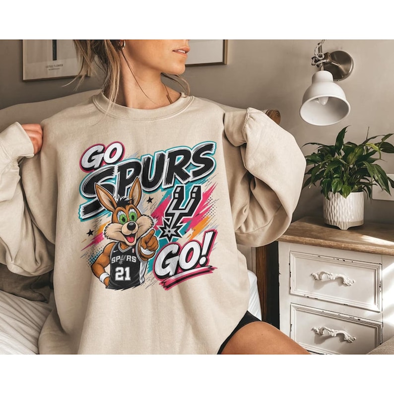 May include: A beige sweatshirt with a graphic featuring the words "GO SPURS GO!" and a cartoon jackrabbit wearing a Spurs jersey with the number 21. The design includes colourful accents and the Spurs logo.