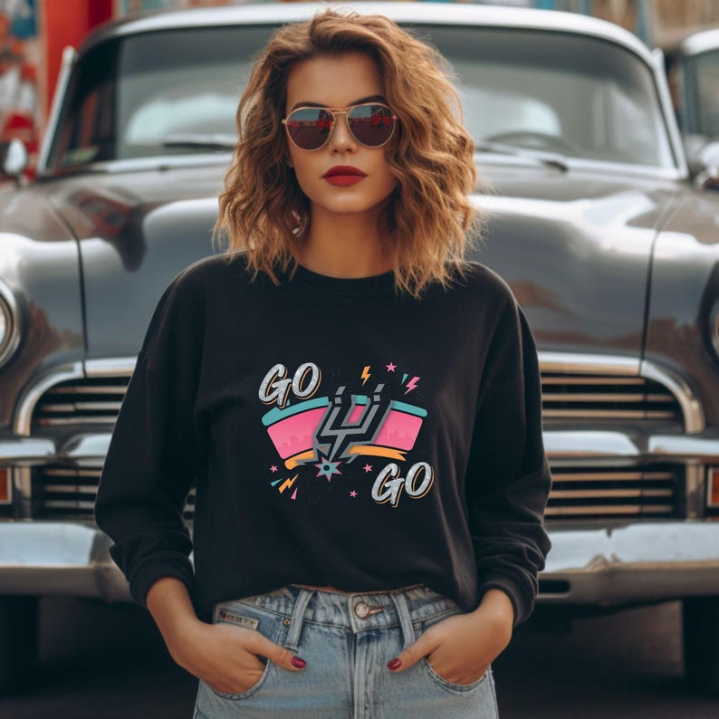 Puede incluir: Sudadera negra de cuello redondo con un dise&ntilde;o gr&aacute;fico colorido con el texto "GO GO" y un emblema estilizado. El dise&ntilde;o incluye elementos rosas, turquesas y amarillos. La persona lleva gafas de sol y vaqueros azules.