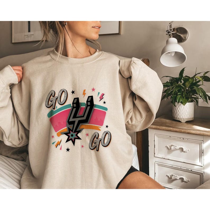 Puede incluir: Sudadera beige con un dise&ntilde;o gr&aacute;fico que presenta las palabras "GO GO" en una fuente estilizada. El dise&ntilde;o incluye una espuela negra y plateada, una estrella y acentos coloridos. La sudadera es de corte holgado.