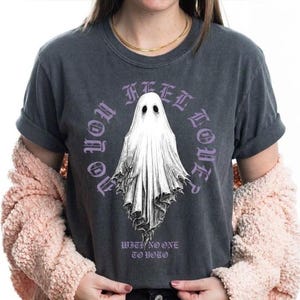 Può includere: T-shirt grigio scuro con un'illustrazione di un fantasma bianco e la scritta viola "NO YOU FEEL ALONE?". Il testo "WITH NO ONE TO YOLO" è sotto il fantasma. La maglietta ha le maniche arrotolate.