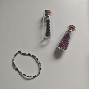Puede incluir: Una pulsera de cuentas con cuentas facetadas de color azul oscuro y magenta alternadas con pequeños espaciadores plateados, acompañada de dos frascos de vidrio transparente con tapones de corcho, uno lleno de cuentas azul oscuro y el otro de cuentas magenta.