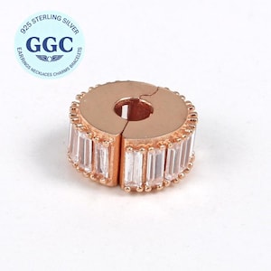 Op de afbeelding: Roségoudkleurige sieradssluiting met een rij rechthoekige, heldere kristallen. De sluiting heeft een cirkelvormige vorm met een gespleten opening. De afbeelding bevat ook een cirkelvormig logo met de tekst "925 STERLING SILVER" en "GGC".