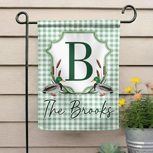 May include: A green and white checkered garden flag with a large green letter "B" in a white framed design. Two duck illustrations flank the letter, with the words "The Brooks" in script below.