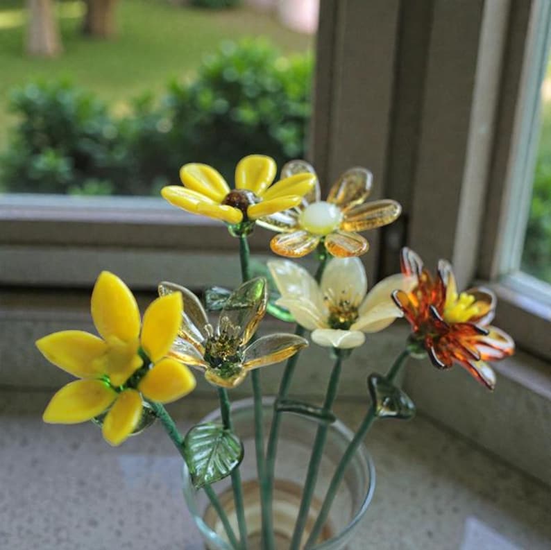 May include: A glass flower arrangement featuring yellow, orange, and clear flowers with green stems and leaves. The flowers are arranged in a clear glass vase, with a blurred background of a window and greenery.