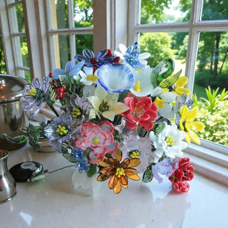 May include: A colorful glass flower bouquet featuring a variety of blooms in shades of red, yellow, blue, and white. The flowers are arranged in a white vase, with a window and greenery in the background.