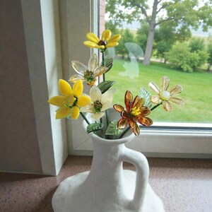 May include: A white ceramic vase with a handle holds a bouquet of glass flowers. The flowers are yellow, white, and amber, with green stems and leaves. The vase sits on a windowsill with a view of a green lawn and trees.