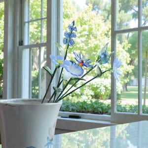 May include: A ceramic vase with a floral design holds a collection of glass flowers. The flowers are primarily blue and white, with green stems and leaves. The vase sits on a table in front of a window.