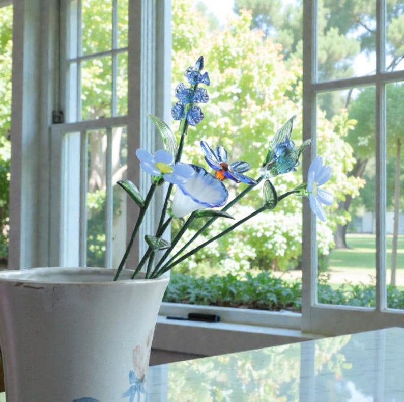 May include: A ceramic vase with a floral design holds a collection of glass flowers. The flowers are primarily blue and white, with green stems and leaves. The vase sits on a table in front of a window.