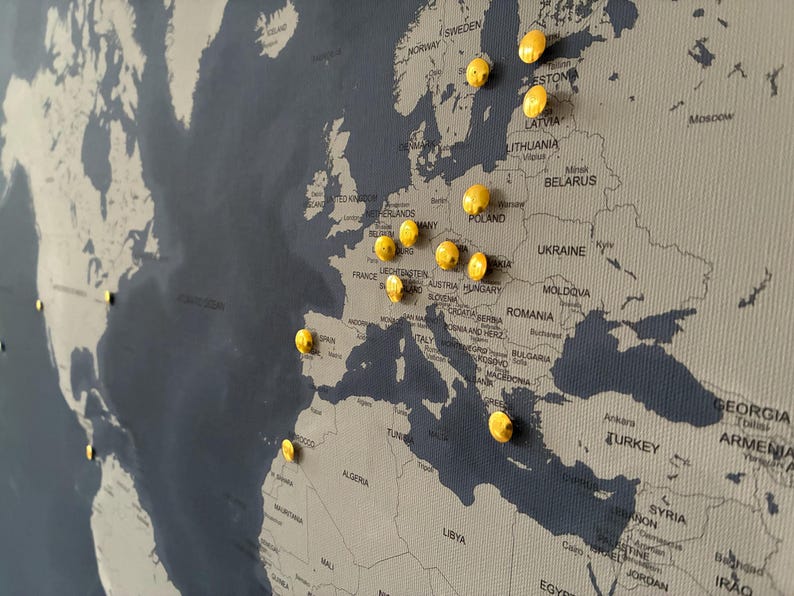 May include: A world map with a dark blue background and light gray continents. Yellow push pins mark various locations in Europe and North America. The map is a decorative item.