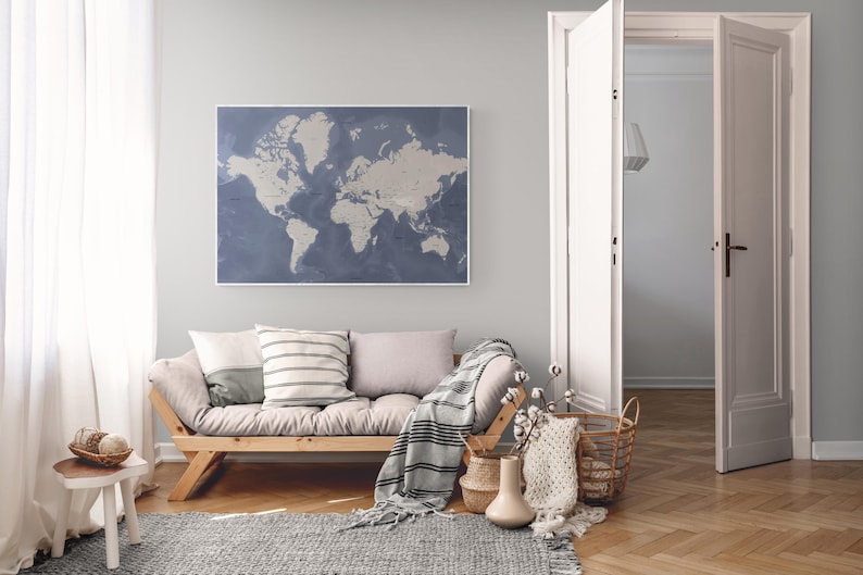 May include: A framed world map in shades of blue and beige hangs above a wooden sofa with pillows and a blanket. The room features a rug, a small side table, and open white doors, creating a cozy and inviting living space. The floor is made of wood.