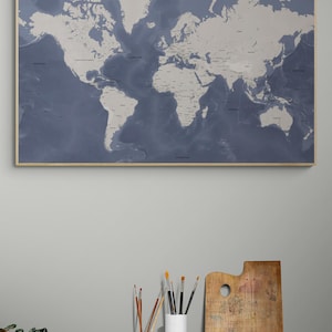 May include: A framed world map in shades of blue and white hangs on a gray wall. The map is detailed, showing continents and countries. Below, a wooden table holds art supplies: paintbrushes in ceramic containers and a wooden palette.