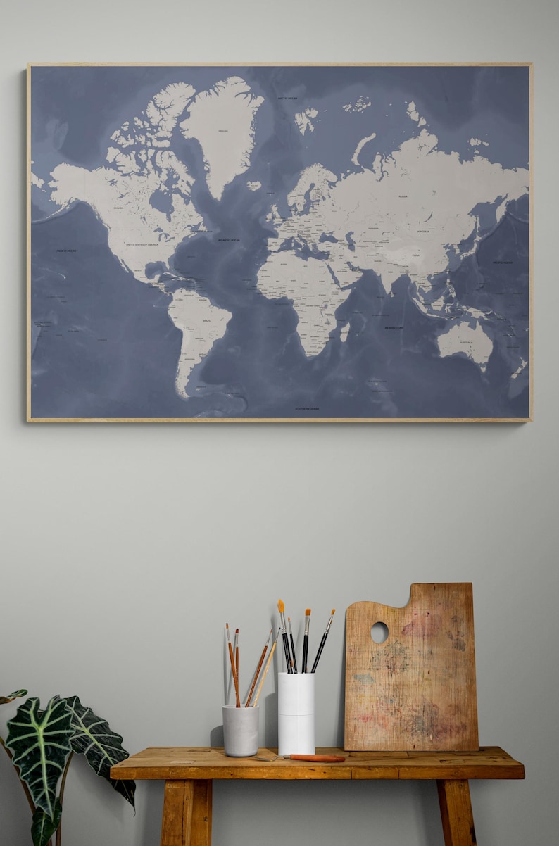 May include: A framed world map in shades of blue and white hangs on a gray wall. The map is detailed, showing continents and countries. Below, a wooden table holds art supplies: paintbrushes in ceramic containers and a wooden palette.