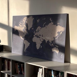 May include: A large world map print with a blue background and beige continents. The map is displayed on a shelf above a bookshelf filled with books. The image is lit by natural sunlight.