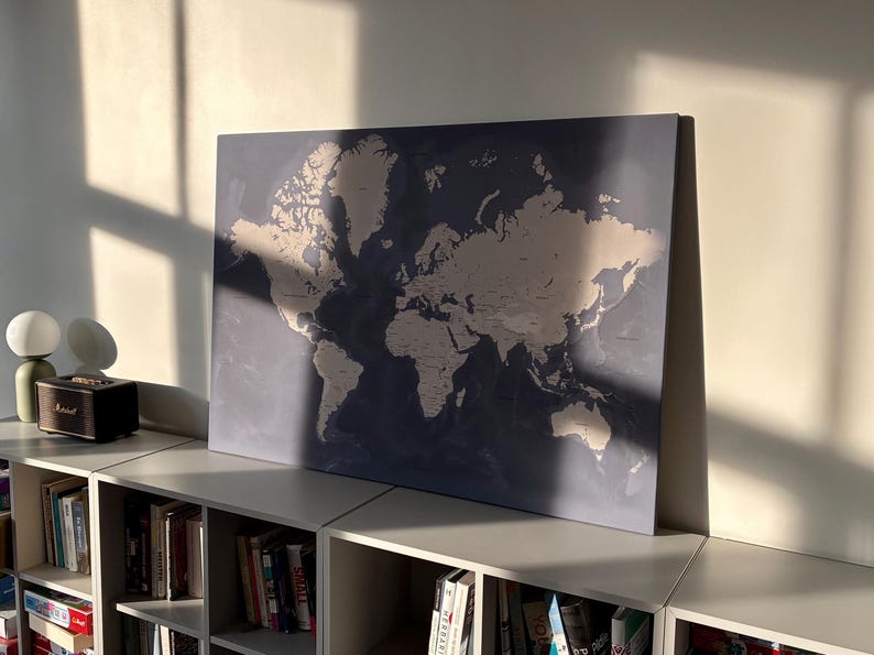 May include: A large world map print with a blue background and beige continents. The map is displayed on a shelf above a bookshelf filled with books. The image is lit by natural sunlight.