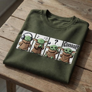 K&ouml;nnte beinhalten: Olivgr&uuml;nes T-Shirt mit einem Comic-Strip-Design. Der Strip zeigt eine gr&uuml;ne Alien-Figur in einem braunen Gewand, mit Kopfh&ouml;rern, Noten, einem Fragezeichen und dem Wort "Coooo!!" in einer Sprechblase.
