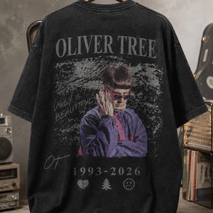 May include: A black acid-wash t-shirt featuring a graphic of Oliver Tree in a purple jacket and red sunglasses. The text reads OLIVER TREE, UGLY IS BEAUTIFUL, and 1993-2026, accompanied by a broken heart, a pine tree, and a sad face icon.