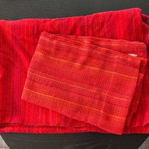 Puede incluir: Un textil rojo doblado con un textil rojo más pequeño y estampado encima. El textil más grande tiene un aspecto texturizado, mientras que el más pequeño presenta rayas horizontales en tonos rojos y naranjas.