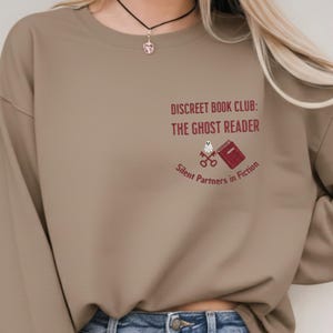 May include: A tan sweatshirt with the text "DISCREET BOOK CLUB: THE GHOST READER" in burgundy. Below the text is a small graphic of a ghost, keys, and a book. The phrase "Silent Partners in Fiction" is below the graphic.