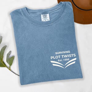 May include: A blue t-shirt with the text "SURVIVING PLOT TWISTS Est. 1998" in white. The shirt is folded and has a crew neck. The Comfort Colors tag is visible at the top. A brown hat is partially visible in the background.