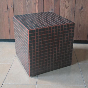 Könnte beinhalten: Würfelförmiger Dekotisch mit Gittermuster. Der Würfel ist hauptsächlich schwarz mit einem roten Gittermuster. Der Tisch besteht aus Holz und steht auf einem gefliesten Boden. Der Hintergrund ist eine Holzwand. Ungefähr 50 cm x 50 cm x 50 cm.