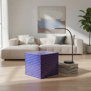 Op de afbeelding: Een blauwe kubusvormige bijzettafel met een rasterpatroon in een woonkamer. De tafel is gemaakt van blauw materiaal met een rasterpatroon in een lichtere kleur. De kamer heeft een witte bank, een zwarte lamp en een plant in een pot.