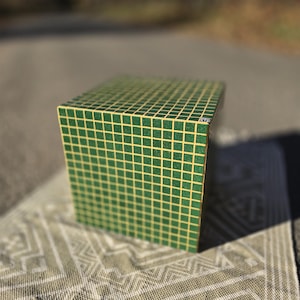 Può includere: Un oggetto a forma di cubo verde con un motivo a griglia di quadrati gialli. L'oggetto è appoggiato su un tappeto beige a fantasia. Il cubo è a fuoco, con uno sfondo sfocato di una strada e alberi.
