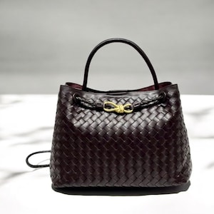 May include: A dark brown woven leather handbag with a top handle and a braided strap detail. The bag features a gold-colored clasp and a small, detachable strap. The interior of the bag is a darker shade of brown.