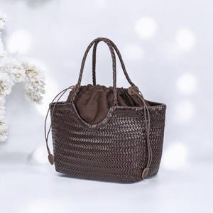 Può includere: Una borsa tote marrone intrecciata con manici intrecciati e chiusura con coulisse. La borsa ha una forma rettangolare con un motivo intrecciato e una fodera in tessuto marrone. I manici sono anch'essi marroni e intrecciati.