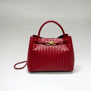 Puede incluir: Un bolso de mano rojo de cuero tejido con un asa superior y un cierre de cordón trenzado. El bolso presenta un cierre de color dorado y una correa de hombro desmontable. El bolso es de tamaño mediano.