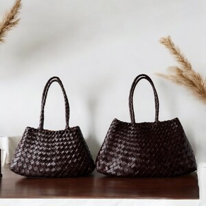 Puede incluir: Dos bolsos tote tejidos de color marrón oscuro con asas trenzadas. Los bolsos tienen forma trapezoidal y se exhiben sobre una superficie de madera. Los bolsos están hechos de un material tejido y las asas están trenzadas. El fondo es una pared blanca lisa.