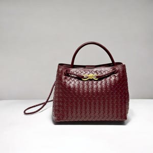 Puede incluir: Bolso de mano de cuero burdeos con diseño tejido. El bolso tiene un asa superior y una correa larga y fina. Un detalle de nudo dorado adorna la parte delantera. El bolso está sobre un fondo neutro.