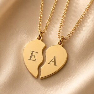 May include: Two gold-coloured necklaces featuring split heart pendants. Each pendant is shaped like one half of a heart and is engraved with a single capital letter, E on the left and A on the right. The pendants hang from delicate gold-coloured chains.