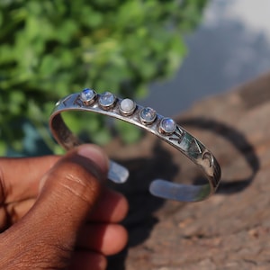Pode incluir: Uma pulseira de punho prateada com cinco cabochões redondos de pedra da lua iridescente. A pulseira tem desenhos geométricos gravados. A pulseira é segurada por uma mão. A pulseira tem aproximadamente 6,35 cm de diâmetro.