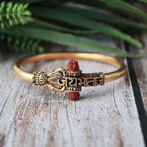 Peut inclure: Bracelet doré avec des détails complexes et un élément décoratif central. Le bracelet présente un bracelet texturé et une pièce maîtresse détaillée avec du texte et une perle brun rougeâtre. Le bracelet est présenté sur une surface en bois.