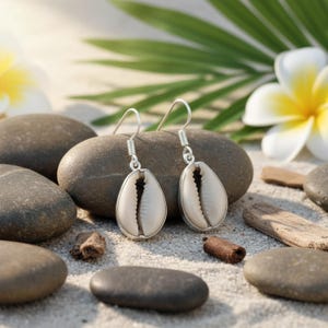 May include: Silver earrings with a teardrop-shaped, off-white seashell design. The earrings are presented on sand with smooth grey stones, driftwood, and tropical flowers in the background.