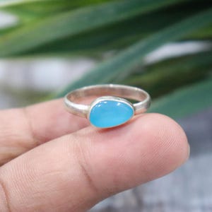 May include: A silver ring featuring a smooth, polished, teardrop-shaped light blue stone set in a simple metal band. The stone has a vibrant, opaque sky-blue color and is securely held within a plain silver bezel setting on a thin, rounded band.