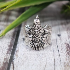 May include: A silver-colored ring featuring a detailed eagle design with outstretched wings. The ring's design includes intricate feather detailing and a prominent eagle head. The ring is set against a wooden background.