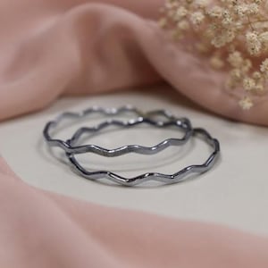 May include: Two thin, metallic gray bangles with a distinct wavy, serpentine silhouette. The surface of each bangle features a fine, textured pattern that catches the light, creating a subtle shimmer across the curved, undulating metal edges.