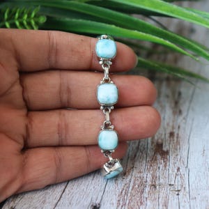 Könnte beinhalten: Ein silbernes Armband mit vier quadratischen, hellblauen Larimar-Steinen. Die Steine sind durch silberne Glieder verbunden. Das Armband wird vor dem Hintergrund von grünem Laub und einer Holzoberfläche gezeigt.