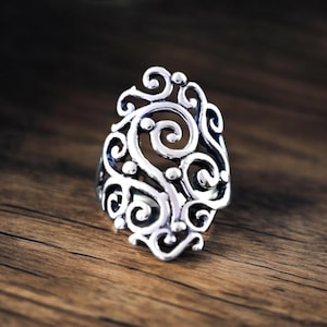May include: A silver-colored ring with an ornate, openwork design. The ring features swirling patterns and small circular accents, set against a dark background. The ring is displayed on a wooden surface.