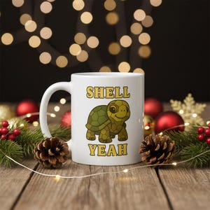 Può includere: Tazza in ceramica bianca con la scritta "SHELL YEAH" e un'illustrazione di una tartaruga dei cartoni animati. La tartaruga ha un guscio verde e una faccia sorridente. La tazza è su una superficie di legno con decorazioni natalizie.