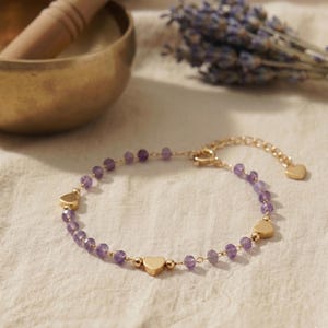 Peut inclure: Un bracelet délicat composé de petites perles d'améthyste violettes à facettes, alternées avec des breloques en forme de cœur en métal doré. Le bijou comprend une chaînette d'extension en métal doré terminée par un petit cœur.