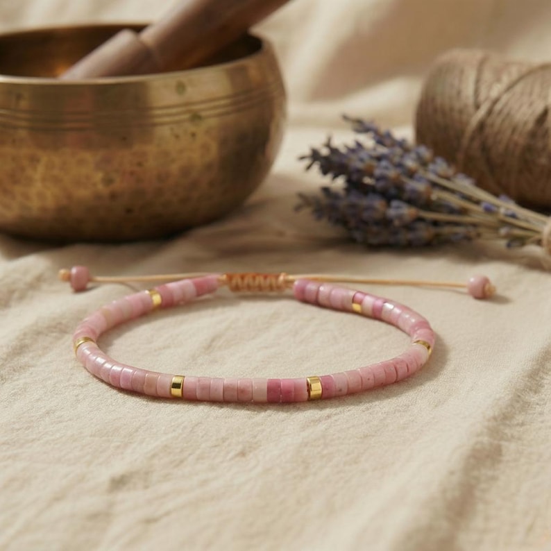 Filigranes Rhodonit Armband aus rosa Natursteinperlen mit goldenen Akzenten auf schwarzer Schieferplatte neben Kerze und Lavendel.