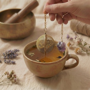 Könnte beinhalten: Ein goldener Tee-Infuser mit einem Mesh-Ball und einem lila Kristallanhänger hängt über einer Keramiktasse mit Tee. Getrocknete Blumen und eine Messingschale befinden sich im Hintergrund und schaffen eine beruhigende Ästhetik.