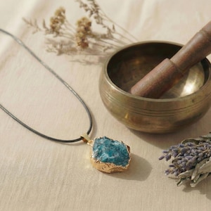 May include: A blue gemstone pendant with a gold-colored bezel hangs from a black cord. A brass singing bowl with a wooden mallet and dried lavender sprigs are in the background. The pendant is a natural, irregular shape.