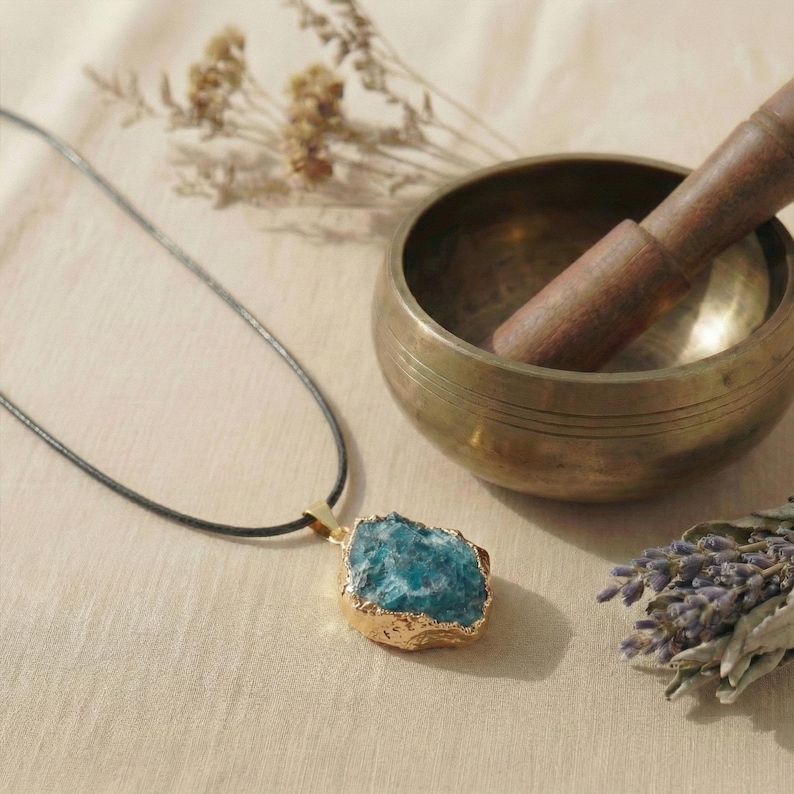 May include: A blue gemstone pendant with a gold-colored bezel hangs from a black cord. A brass singing bowl with a wooden mallet and dried lavender sprigs are in the background. The pendant is a natural, irregular shape.