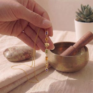 Hand hält den langen vergoldeten 7-Chakra-Anhänger senkrecht neben einer tibetischen Klangschale.
