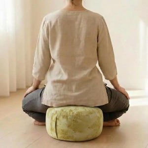 Puede incluir: Una persona está sentada sobre un cojín de meditación redondo verde claro. El cojín tiene un diseño plisado y una apariencia texturizada. La persona lleva una parte superior beige con mangas tres cuartos y pantalones grises. El entorno parece ser interior.