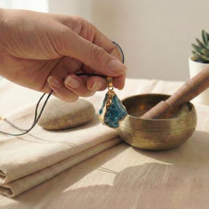 May include: A blue gemstone pendant with gold-colored trim, suspended from a black cord. The pendant is held in a hand, with a brass bowl and wooden pestle in the background. A small rock and folded cloth are also visible.